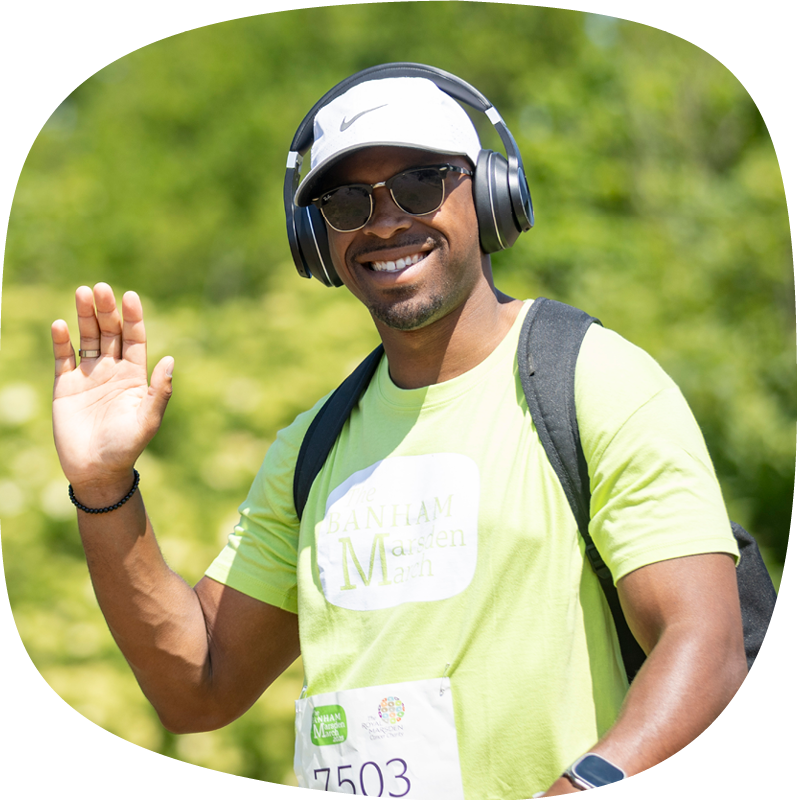 A Banham Marsden Marcher smiling and waving. They're wearing the green Banham Marsden March t-shirt, a white cap and black over-the-ear headphones.