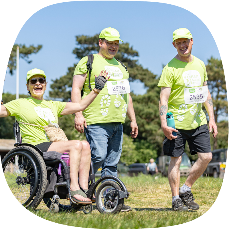 Three people taking part in the Banham Marsden March. One person is in a wheelchair, smiling with their hands up. The other two people are smiling and walking alongside