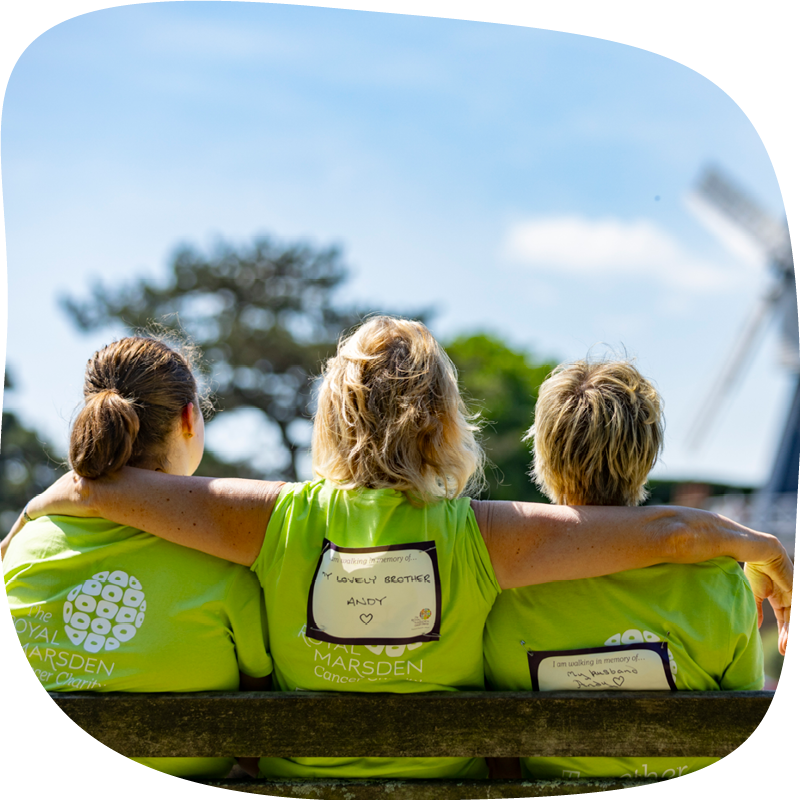 The backs of three Banham Marsden Marchers sitting on a bench in a park. The middle person has their arms around the two people either side. In front of them are trees and a windmill.