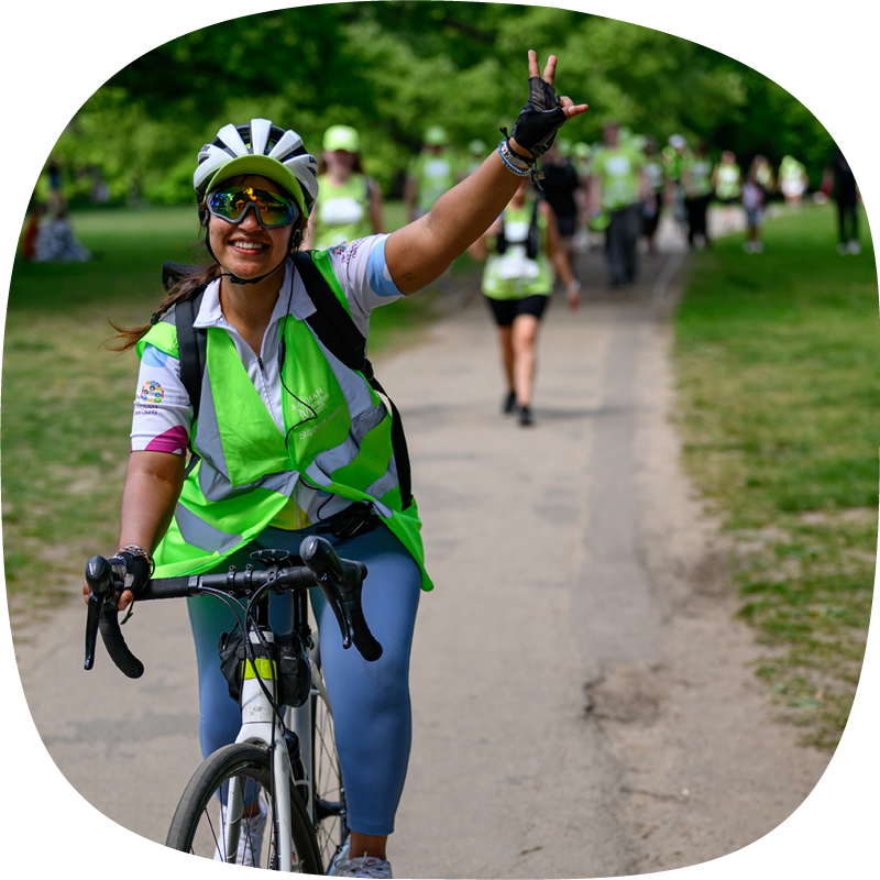 A Banham Marsden March volunteer smiling and riding a bike, holding the handlebar with one hand and giving a peace sign with the other. They're wearing a high-vis jacket, helmet and sunglasses